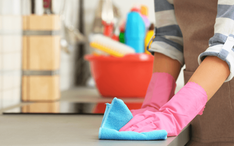 Learn To Keep Your Kitchen Neat And Tidy With These Cleaning Tips Image of a hand wiping a kitchen counter using a cleaning rag, with a basket of cleaning supplies in the background (spray bottles, rags, etc.)