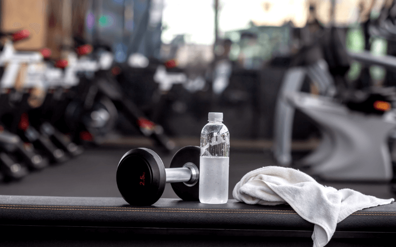 8 Dumbbell Exercises For An Effective At-Home Workout Routine Image of a workout bench with a pair of dumbbells on top, plus a water bottle