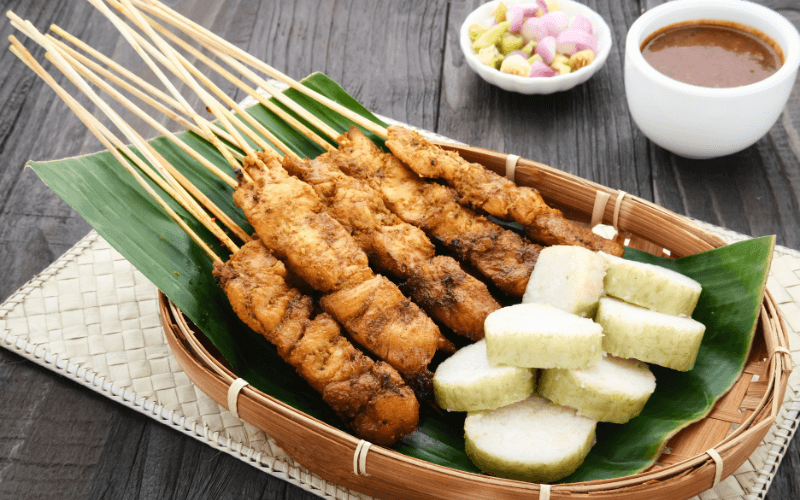 Image shot of a plate of Satay, traditionally served on a banana leaf with a side of sauce, surrounded by other dishes.